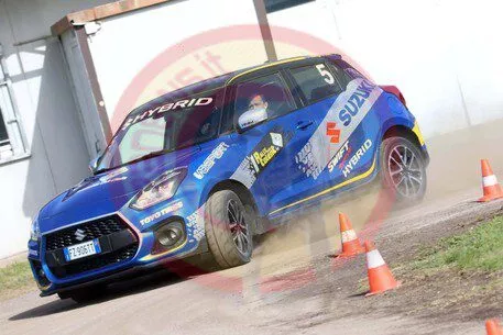 Un'auto da rally Suzuki Swift Hybrid blu, la numero 5, sfreccia su una pista sterrata all'aperto, sollevando polvere e attraversando coni stradali arancioni. L'auto è ricoperta di loghi e decalcomanie degli sponsor.