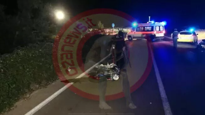 Scena stradale notturna dopo un incidente, con veicoli di emergenza, luci lampeggianti e soccorritori; una persona in primo piano tiene in mano parti di bicicletta danneggiate.