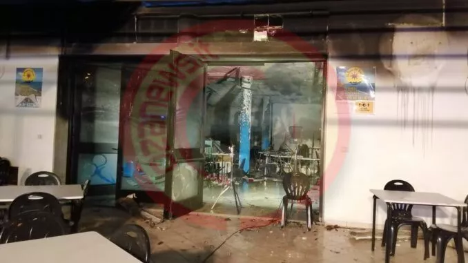 Un bar danneggiato con pareti annerite e detriti sparsi all'esterno. Sedie e tavoli di plastica sono sistemati vicino all'ingresso, che presenta vetri in frantumi e danni visibili da incendio all'interno. Alle pareti sono appesi dei manifesti.